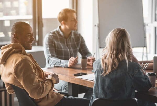Group Voluntary Insurance - Four Young People Sitting at an Office Conference Table Together and Conversing and Laughing with Coffee Cups on the Desk